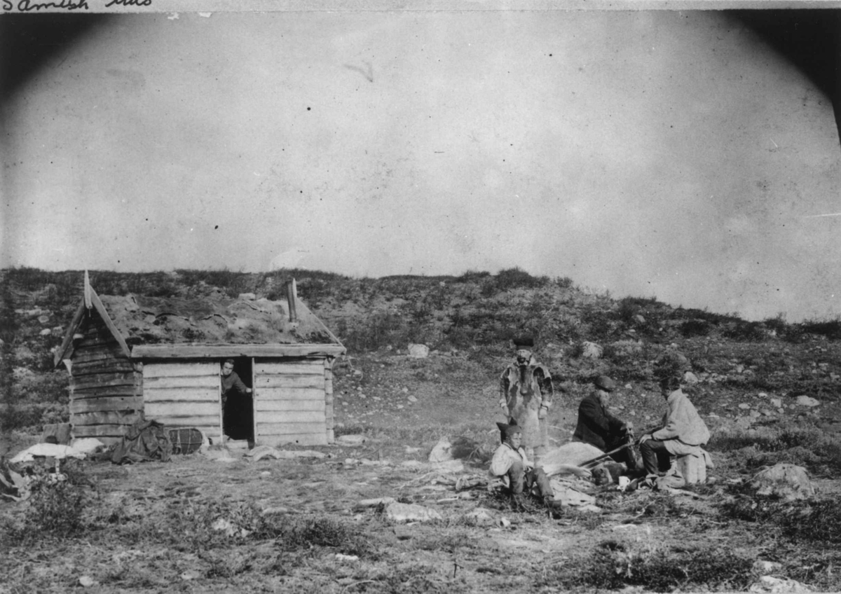 Tømret hus med en person i døråpningen, fire peroner utenfor. Finnmark 1897.