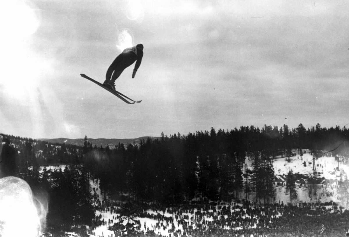 Ski. Hopprenn. Holmenkollen, Oslo 1934. En hopper i svevet. Publikum ses på sletta. Nordmarka i bakgrunnen