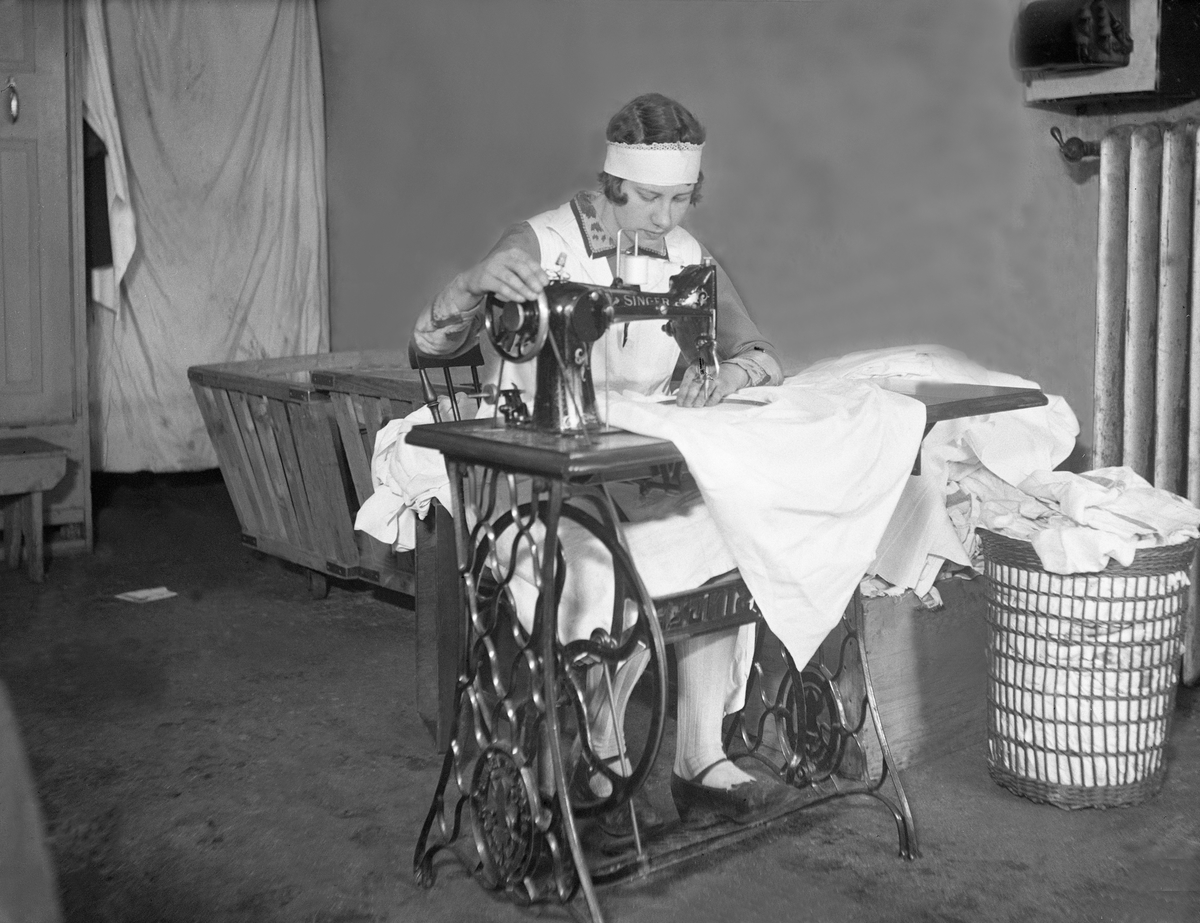 En kvinnelig ansatt i Vaskeri A/S i Oslo sitter ved en symaskin å reparere tøy, Fotografert ca. 1929.