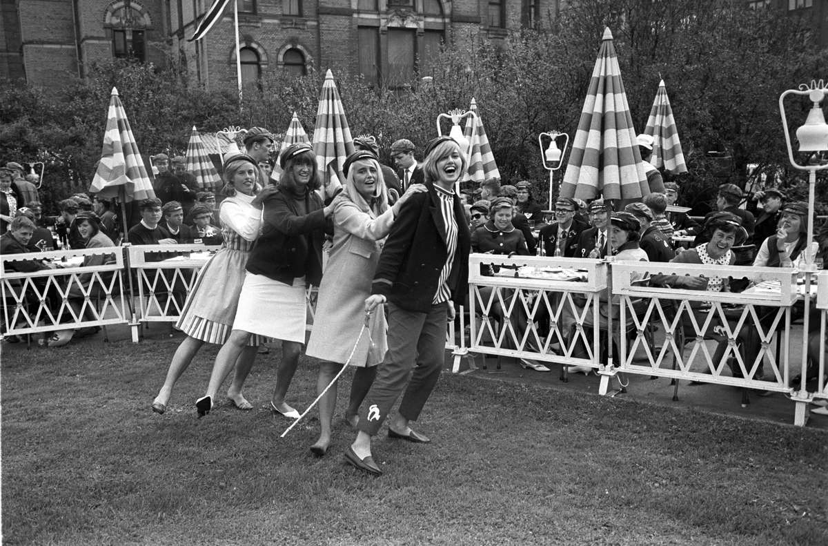 Russen danser jenka og viser russemoter på gressplenen utenfor uterestauranten Pernille i Oslo i mai 1965. Antatt russeprinsesse danser først i rekka. Nationaltheatret skimtes i bakgrunnen.
