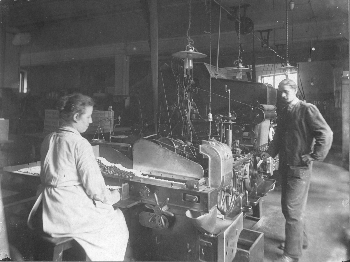 Interiørbilde fra tobakksfabrikken W. Hartog & Co. En kvinne og en mann arbeider ved en sigarettmaskin.
