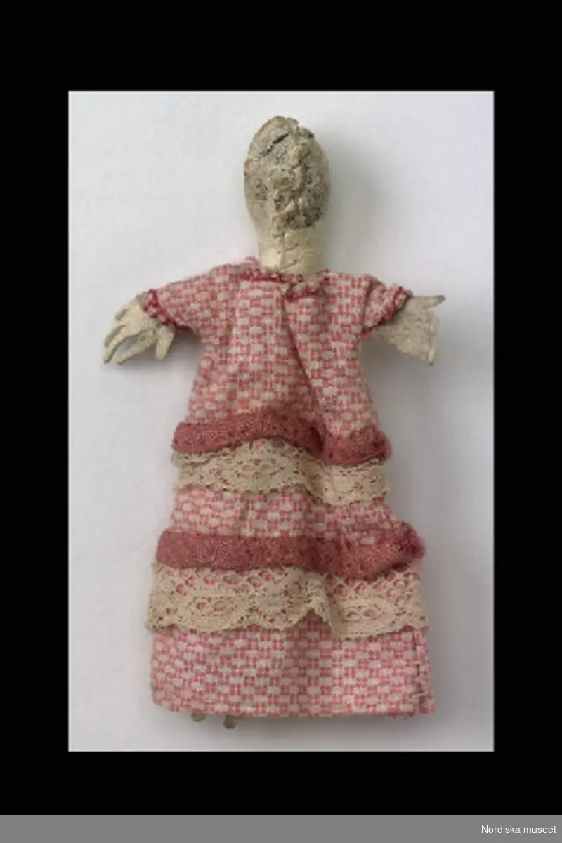 Inventering Sesam 1996-1999:
L  7,5  cm
Docka, hemsydd, kropp av vitt skinn, rosa bomulls klänning. 
Dockan tillhör en samling leksaker 108.216-108.456.
Birgitta Martinius 1996