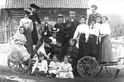 Gruppeportrett, gårdsfolk og besøkende samlet ved vogn på tu