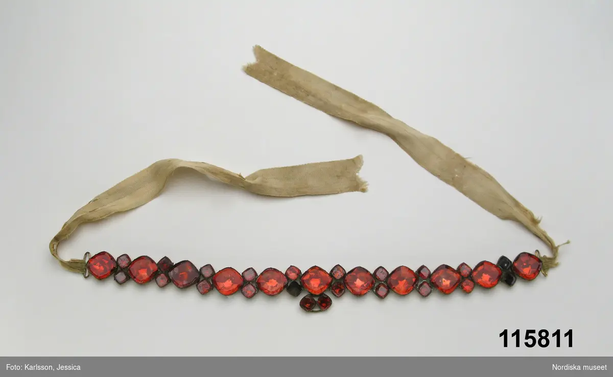 Huvudliggaren:
"Halsband av större och mindre fyrsidiga, röda glasbitar, infattade i förgylld mänssing. 1700-talet."