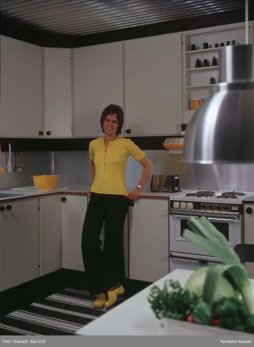 Kvinna klädd i gul jumper och gula träskor, lutar sig mot arbetsbänk i kök. På bänken en gul plastbunke.