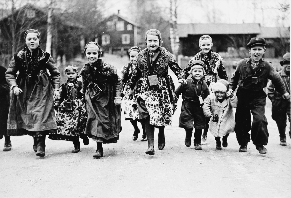 Barn, små kullor och masar, i folkdräkt springer på väg mot fotograf. Barnen kallar till Valborgsmässa 30 april 1936 i Floda genom att ringa i koskällor.