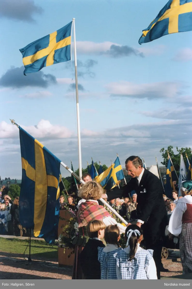 Sveriges Nationaldagsfirande på Skansen, Svenska Flaggans dag. Kung Carl XVI Gustav delar ut en fana till kvinna folkdräkt.