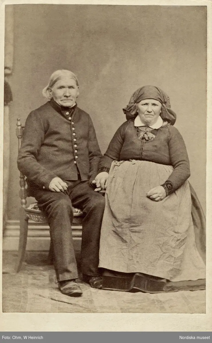 Porträtt av Häradsdomaren Johan Nilsson i Örup och hans maka Inger. Bara hd. Skåne. Paret sitter på varsin stol och håller varndra i handen.