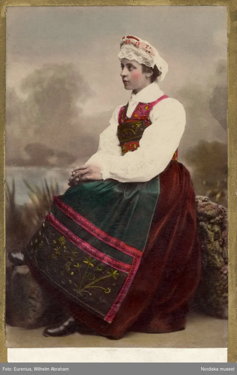 En kvinna i folkdräkt sitter vänd i profil. Högtidsdräkt från Österåker, Södermanland. Som den bars omkr. sekelskiftet 1900
