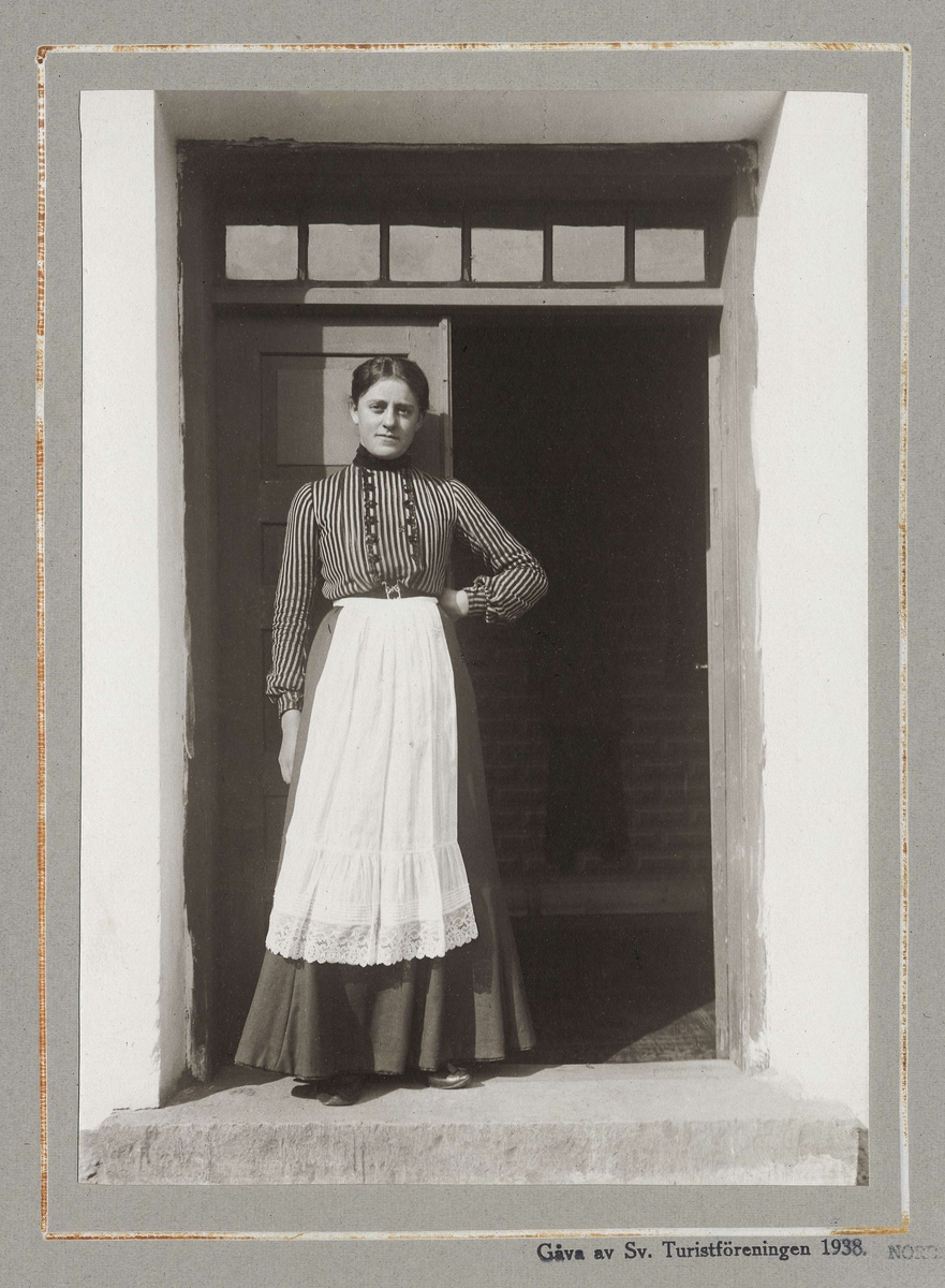 Ung kvinna med förkläde i dörröppning. Bonddotter från Västra Klagstorp, Skåne.