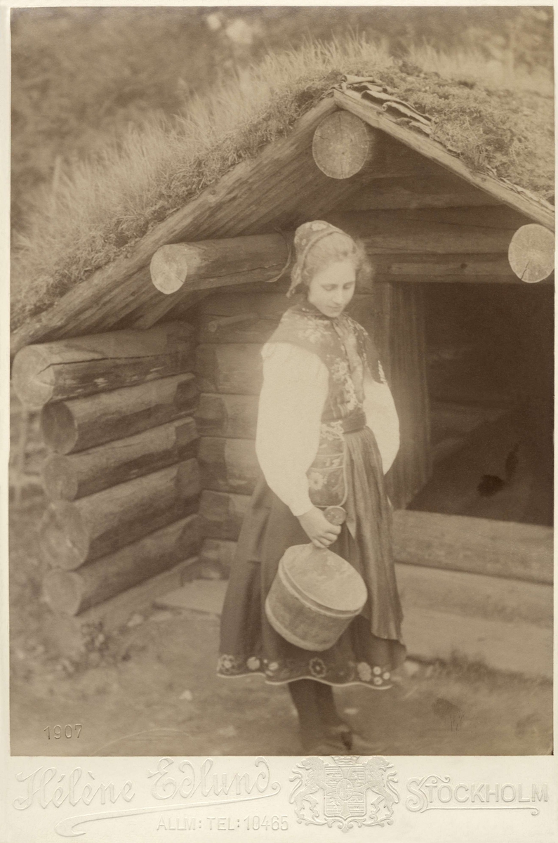 En ung kvinna står utanför ett timrat hus, med en träbytta i handen