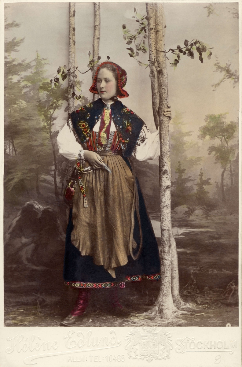 Kvinna i helfigur klädd i dräkt från Floda socken, Dalarna. Handkolorerat fotografi