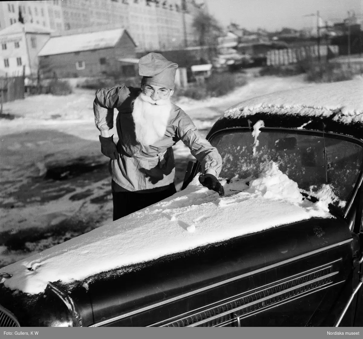 Man i jultomtedräkt borstar bort snö från bil.