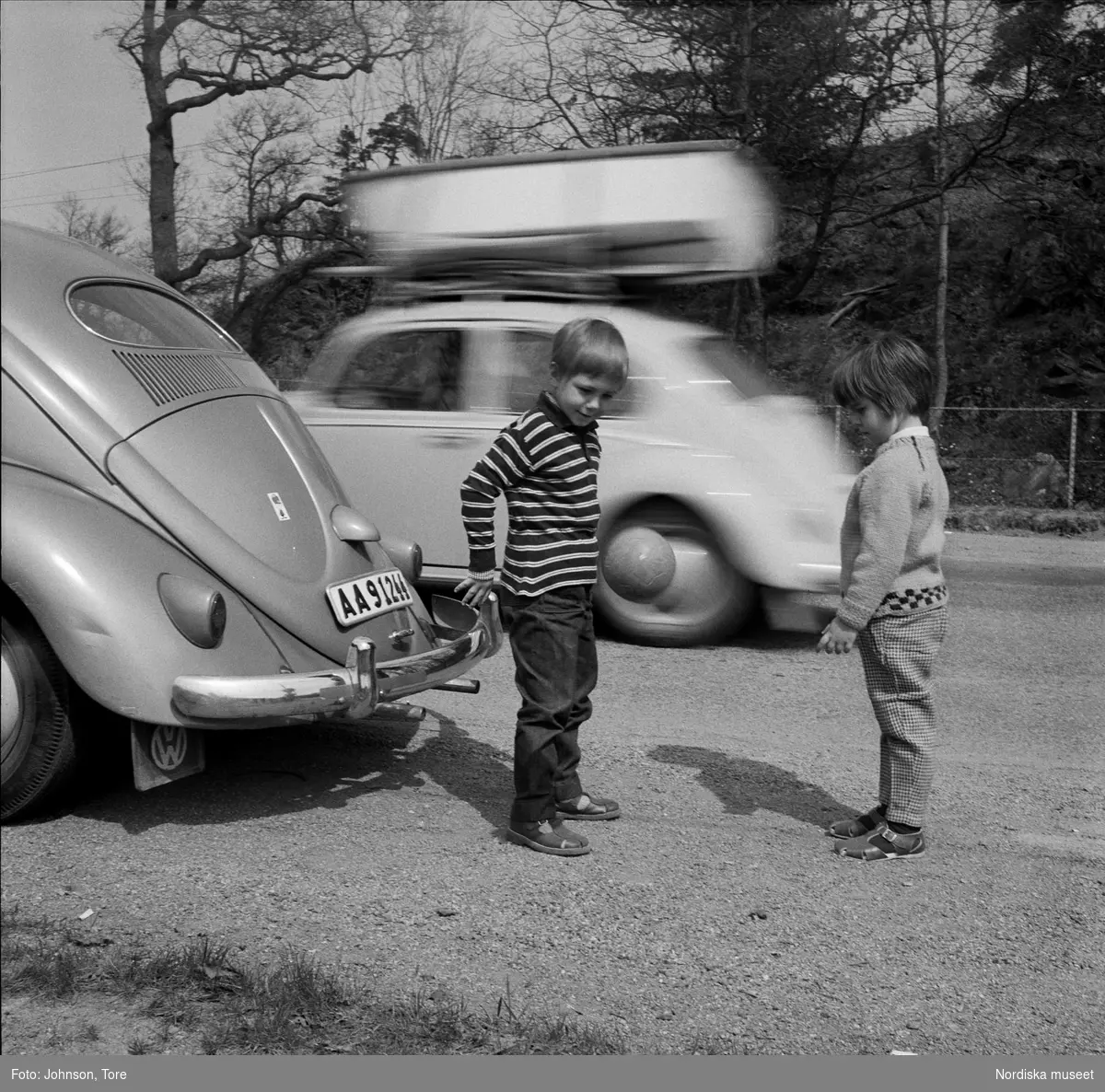 "Barn i trafik". Två barn leker med boll vid vägkant, passerande bil. Parkerad bild, Volkswagen.