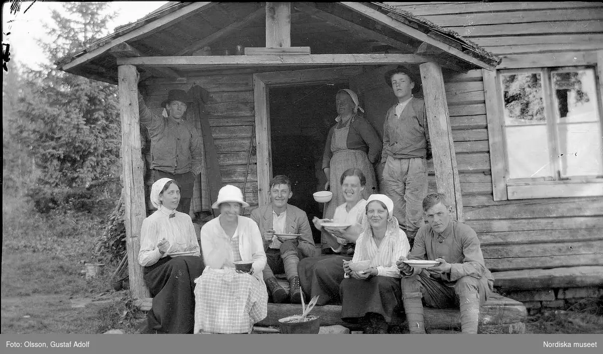 En grupp män och kvinnor sitter och äter på trappan till ett hus. 1900-talets första hälft.