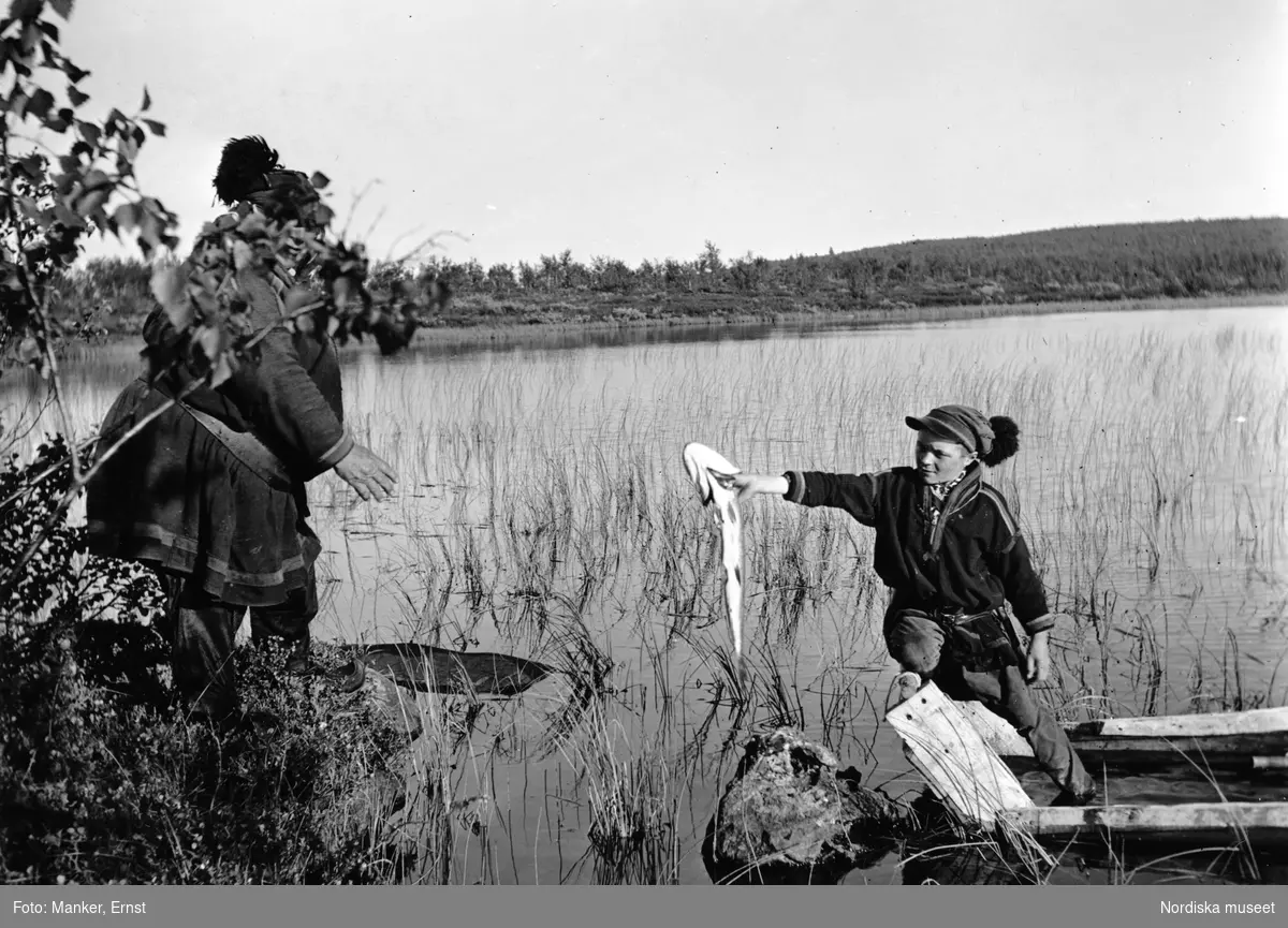 Pskrift: "Nils Nilsson Skum med sin fosterson Nikolaus Huvva på en fisketur. Nikolaus räcker i land en gädda, som han drivit mot ett nät i vassen. En mindre sjö norr om Sjisjkavare." Lappland, Gällivare socken, Norrkaitum, Sjisjkavare.