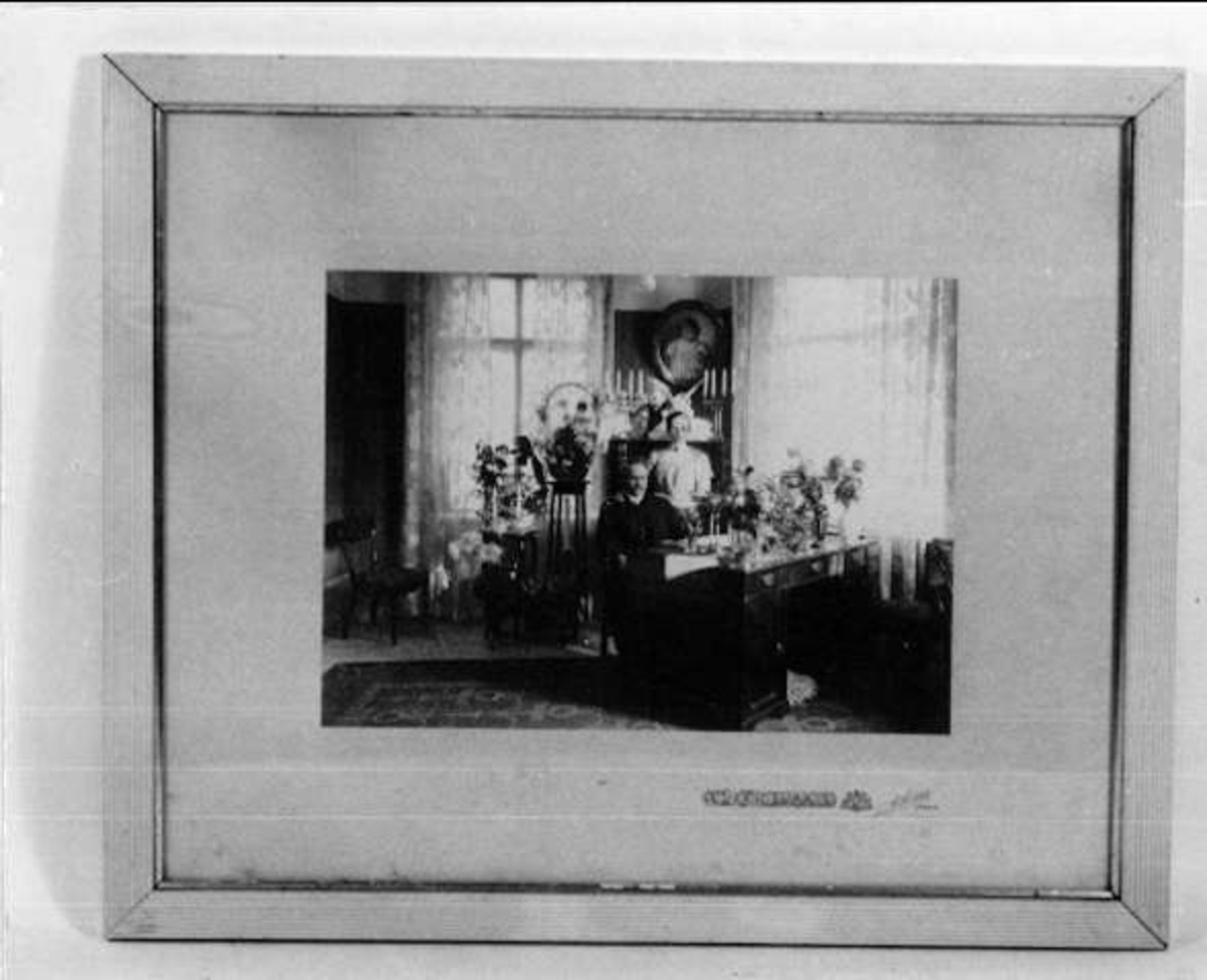 Svartvitt fotografi, interiör med en man, Hjalmar Söderberg, grundare av  AB Hjalmar Söderberg. Han sitter vid skrivbordet och en kvinna står bredvid. Blommor på bordet och runt bordet.