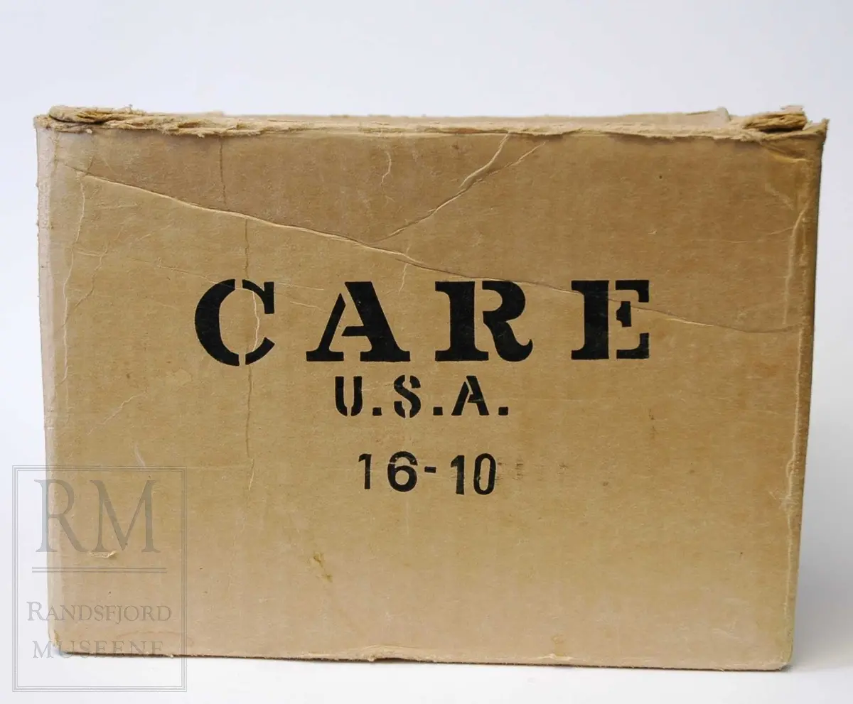 Kvadratisk, avlang form. Pappeske for sending med varer til kriserammede/behovstrengende områder.
Innhold i esken: To pakker med ullstoff. Det ene er i mørk/lys beige: Dreil/ fiskebensvevd kyper. Det andre i ensfarvet brun, samme teknikk.

