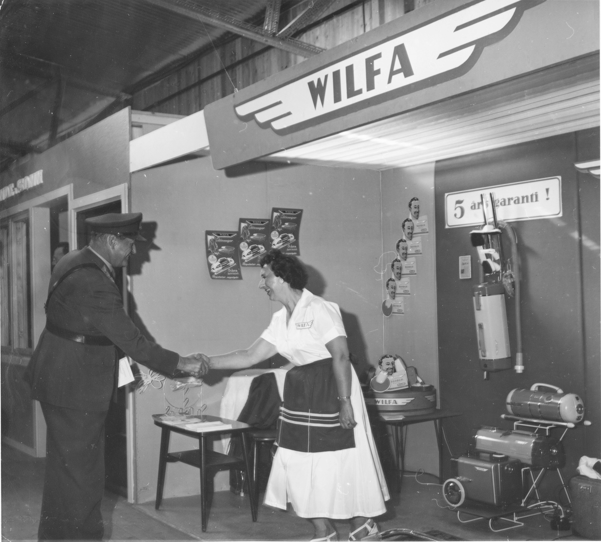 Kong Olav hilser på en dame som står på Wilfa sin stand i forbindelse med en firmautstilling / varerepresentasjon.