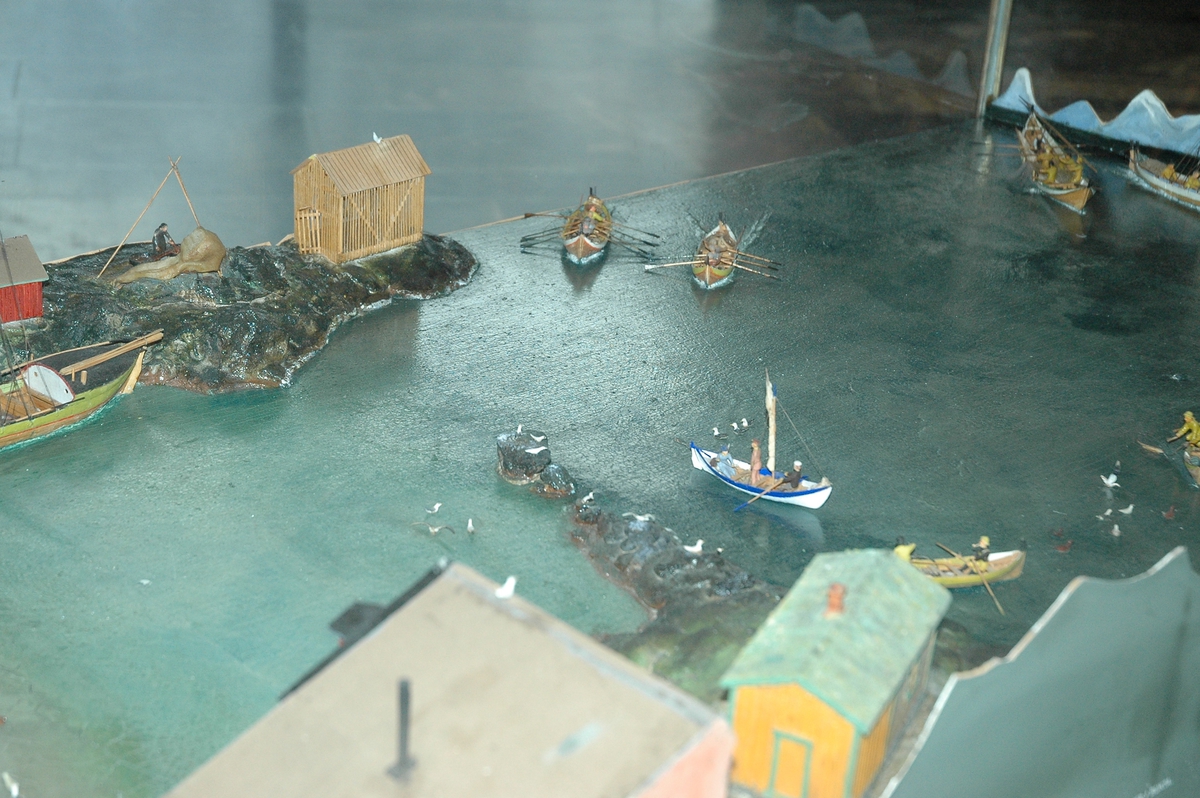 Viser fiskevær i Bø i Vesterålen, glasskasse med stålunderstell. Viser landskap med sjø, båter og hus, noe menneskelig aktivitet.