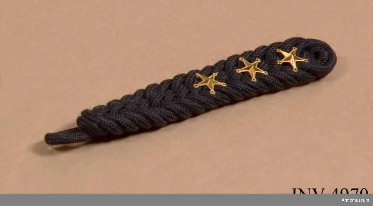 Fläta av svart yllegarn (svart trådflätning) m gradbeteckning i form av tre guldstjärnor m/1858. Används t kollett.