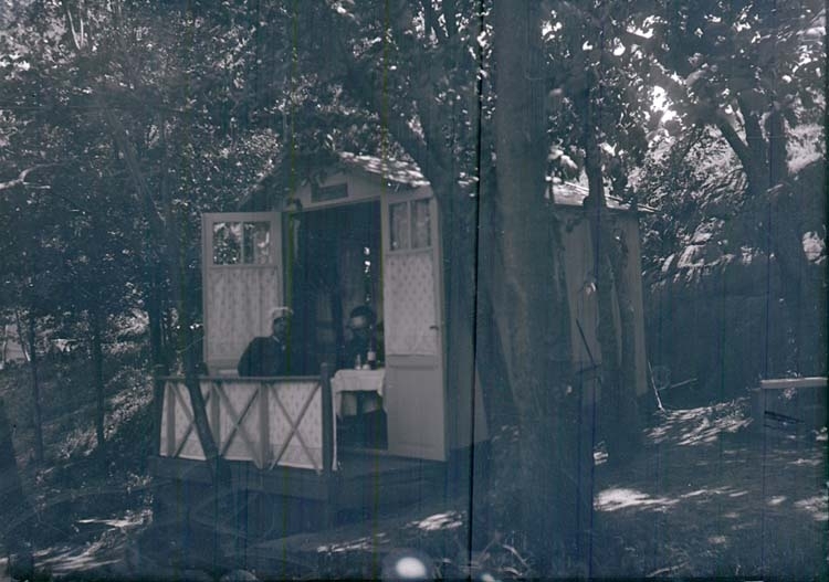 G.L. Forslind och en annan man sitter i ett litet hus i en trädgård.
"G.L. Forslind vid Samskolan 1915 L.l."