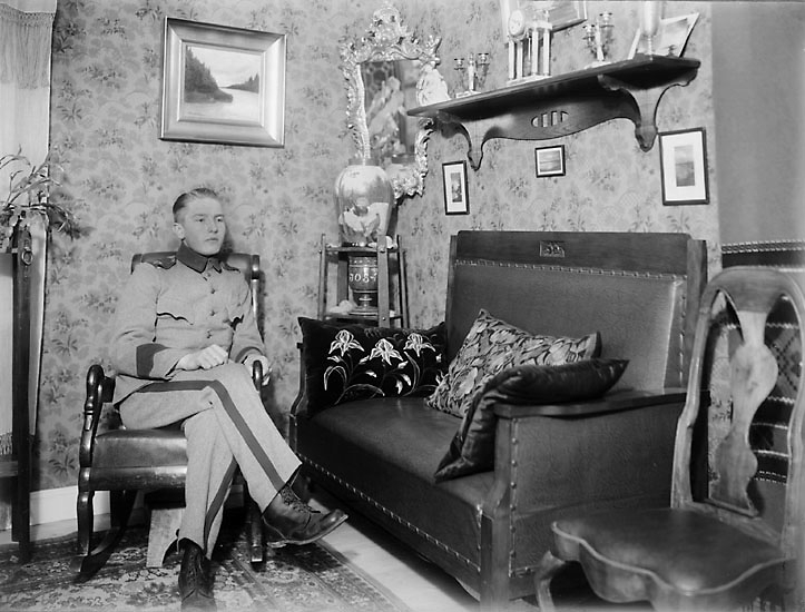 En man i uniform (m/10) sitter i en gungstol. Bredvid honom står en soffa med broderade kuddar. I hörnet hänger en spegel med förgylld ram.