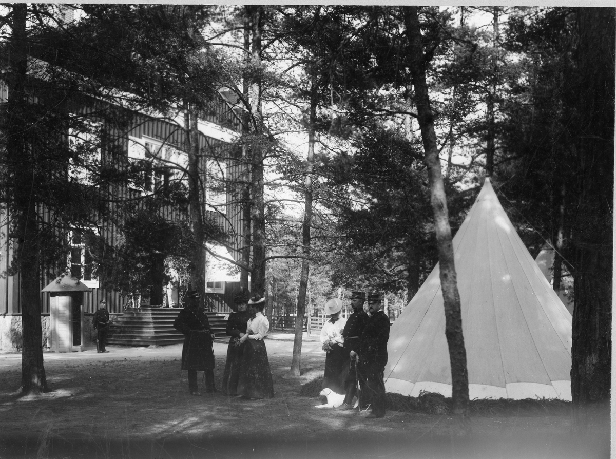 Soldater vid Hälsinge regemente I 14, med kvinnligt sällskap utanför tält framför byggnad med bemannad vaktkur.