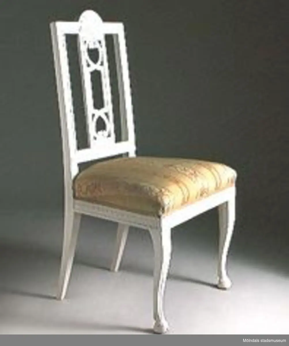 Stol, sengustaviansk, med s.k. "Gunnebobricka" i ryggen. Solfjädersornament på krön och bladsnitt i rygg och sarg. Frambenen med fotavslutningar i form av skurna bockhovar. Stolen har ursprungligen varit målad i en brunröd nyans. Vitmålad på 1900-talet.Halva stolen med framtagen originalfärg. Stolen är osignerad men är sannolikt tillverkad i Lindome omkring år 1800. Ryggbrickans genombrutna utformning återgår på en ritning av Carl Wilhelm Carlberg för en stol Gunnebo, därav namnet "Gunnebobricka". Denna stol en utomordentligt hög kvalitet i skärningarna och benens utformning. Troligen är stolen del av ett större möblemang och utfört som en specialbeställning.Inköpt från antikhandeln Stopalo, Stockholm.