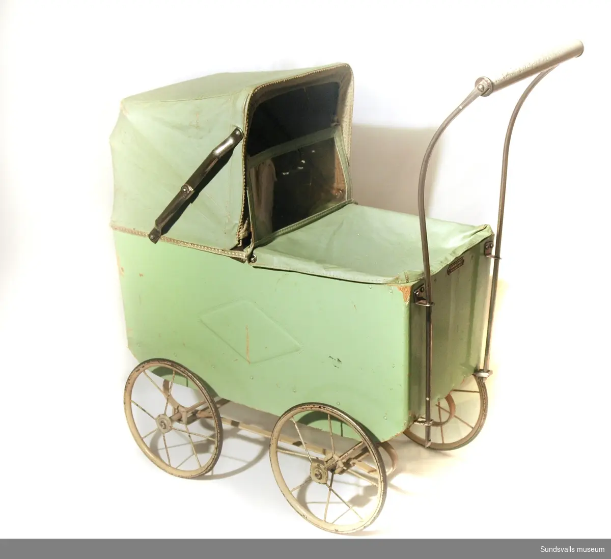 Dockvagn i ljusgrönt, med vagn av trä och suflett av vaxduk. På vagnens sida finns ett mönster i form av en fyrkant. I vagnen ligger även en virkad filt.