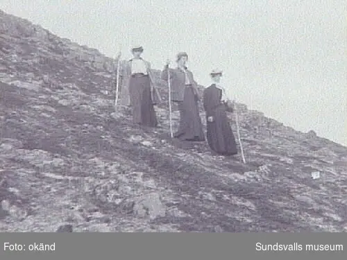 Tre kvinnor med vandringsstavar och långa kjolar, går nedför bergsluttning.