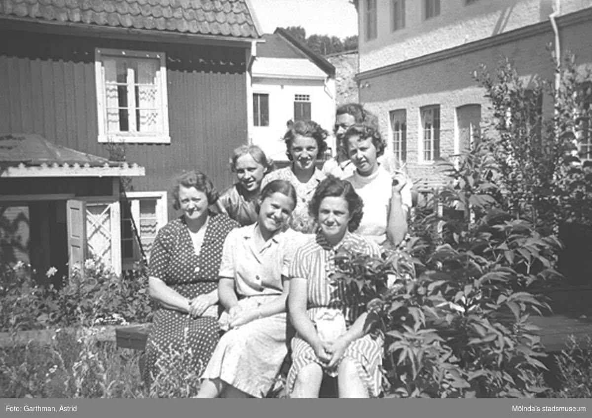 Sju kvinnliga arbetskamrater (okända för givaren) står/sitter i gräset framför ett hus och en fabrik. De är från "Lumpans" (Forsåkers fabriker), Kvarnfallet 31. Detta är öster om järnvägsvallen under 1950-talet.