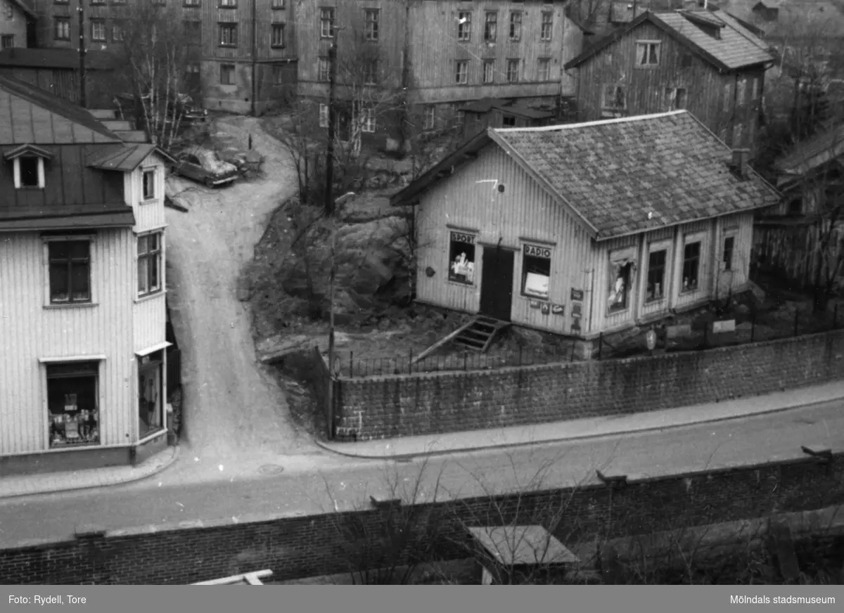 Vy från pappersbruket Papyrus område i Forsåker, Mölndal, mot bebyggelse vid Kvarnbygatan i Mölndals Kvarnby på 1960-talet. Till vänster ses nr 27 som inrymde C.J. Johanssons Järnhandel. Till höger ses nr 29 som var Mölndals första stadshus. En rådstuga som med tiden blev affär för radio och sport.