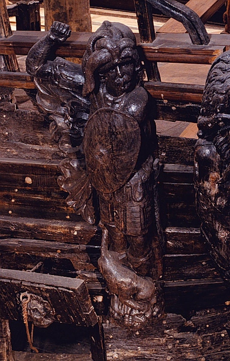 Skulptur föreställande en högrest krigare med stor, tredelad mustasch. Mannen är iklädd en romersk rustning av thoraxmodell och på huvudet sitter en slät hjälm med långa vajande plymer. Vid hans högra sida syns en veckad mantel som fladdrar i vinden. På fötterna syns en sandalliknande fotbeklädnad som sträcker sig upp på smalbenen.

Med sin vänstra hand håller krigaren en stor, oval sköld tryckt tätt mot bröstet. Skölden är prydd med symmetriskt anbragta, vågformiga räfflingar. Mitt på skölden finns en rundad upphöjning med ett hål där troligtivs en sköldprydnad, i form av en sköldbuckla eller dylikt, suttit fästad.



Hans högra arm hålls lyftad rakt ut från sidan och underarmen är böjd i en vinkel uppåt. I sin knutna hand har han hållit ett böjt svärd (eller kroksabel eller sabel) som han har dragit från svärdsskidan hängande vid hans vänstra ben. Huvudet är vridet mot vänster. Kroppstyngden vilar på höger ben medan vänster ben hålls något böjt.

Krigaren står på ett stort, frontalställt lejonhuvud. Lejonets framtassar är synliga. Framför mannens fötter, ovanpå lejonhuvudet, sitter en liten hund. Hunden är placerad diagonalt och ses i profil. Hunden har stora öron, lång, spetsig nos och lång, kraftig svans.

Figuren är skuren i ett stycke tillsammans med en stor ryggpanel vilkens baksida med urtag indikerar att skulpturen har suttit på skeppets utsida, på bordläggningen. Höger arm, manteln och svärdsskidan är dock snidade separat.

Skulpturen är mycket välbevarad. Hundens svans är bruten.



Text in English: A sculpture of a lofty warrior clad in Roman armour of the thorax type, wearing a plain helmet with long, wavy plumes. The man has a three-lobed moustache but is otherwise clean-shaven. He wears a sandal-like footwear that extend up to his shins. To the right a cloak gathered into folds flaps in the wind. In his left hand he holds a large oval shield tightly against his breast: the shield is embellished with symmetrically applied wavelike grooves and has a rounded protrusion in the middle with a hole in to which some form of a shield decoration may have been attached.

The right arm is held out from the body, at an angle, with the hand clenched, possibly having held a sword (or a scimitar or sabre) which he has withdrawn from the scabbard hanging by his left leg. The warrior has his head turned to the left, and he rests on his right leg while the left leg is somewhat bent.

The figure stands on a lion''s head facing forwards with its forepaws visible. In front of the man''s feet, and on the lion''s head, there is a small dog seen in profile, with long pointed nose, large drooping ears and a powerful tail. The figure is carved in one piece except for the right arm, the cloak and the scabbard which have been added separately. It has a large back-board of which the back reveals that the sculpture was attached to the outside planking of the ship.

The sculpture is particularly well preserved, with several traces of gold visible at the time of the salvaging. The dog''s tail is broken.