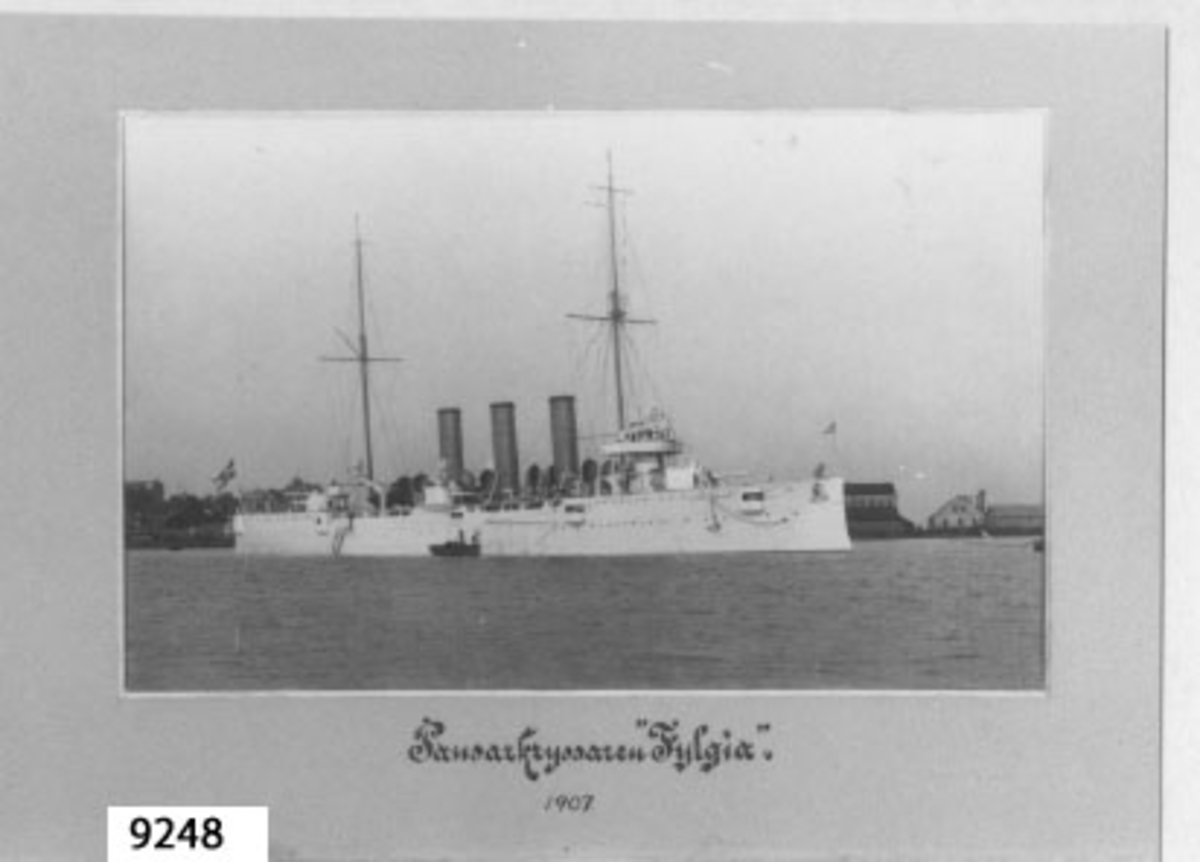Fotografi inom ram av papp, ljusgrå.
Visar pansarkryssaren FYLGIA förtöjd på Karlskrona örlolgsvarv år 1907.
Text i svart: Pansarkryssaren Fylgia.