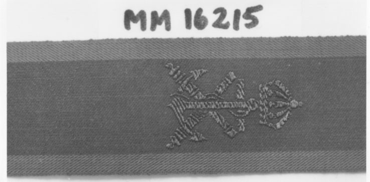 Vapenslagsbeteckning, textilband med tre gröna längsgående fält: märkning för vikning. Vapenslagstecken för Marinen, två korslagda kanonrör med på sig ett ankare krönt med krona: invävt i bronsfärgad tråd.