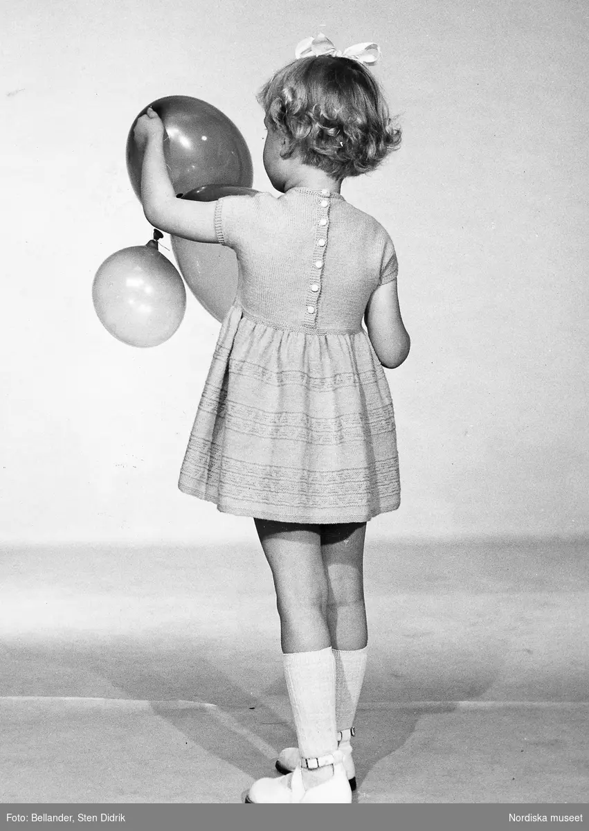 Ett barn i klänning, rosett, knästrumpor och sandaler håller i ballonger.