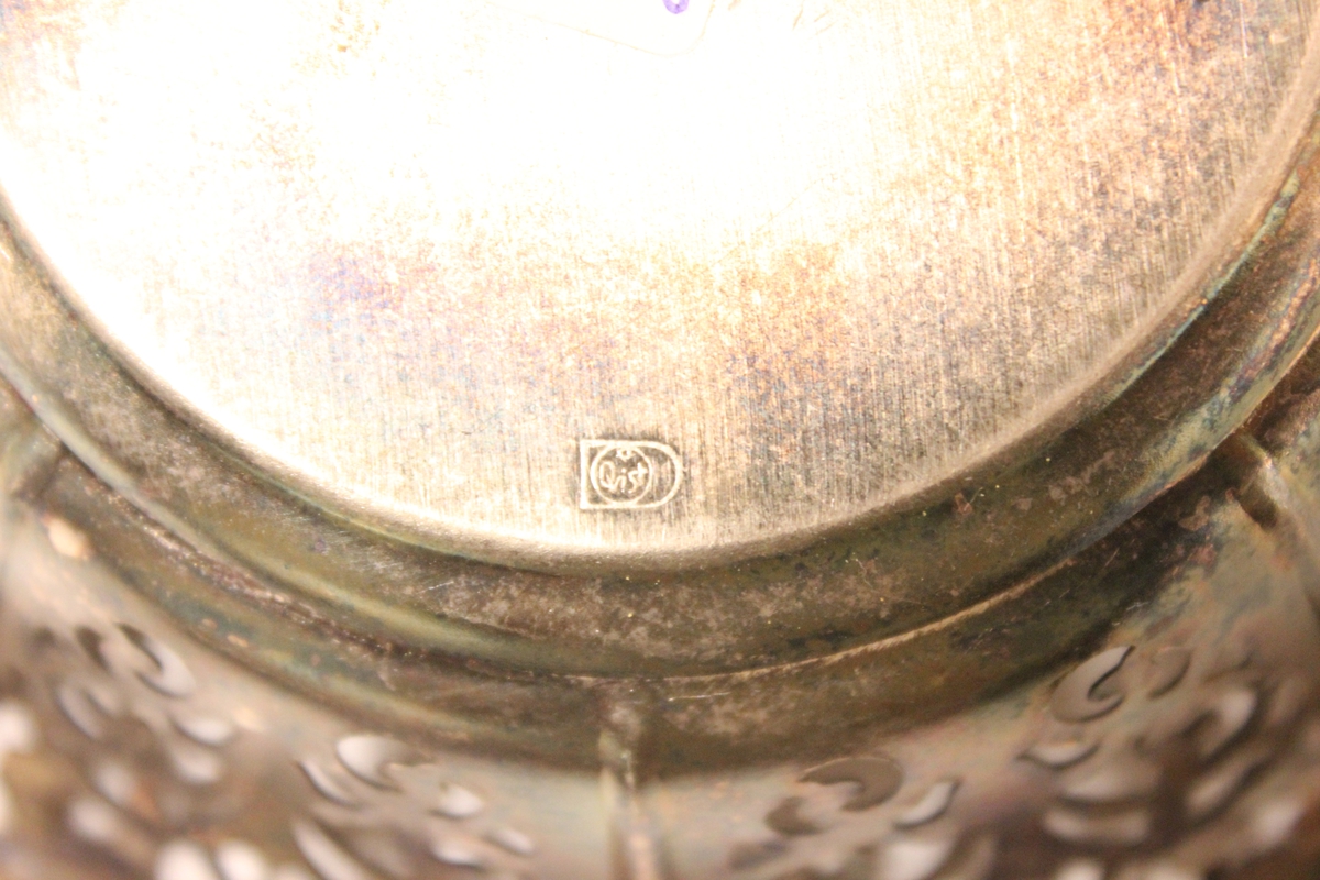 En lite skål i sølplett med mønstret hullbård rundt hele skålen. Under skålen er det et preget stempel som det står "Quist"