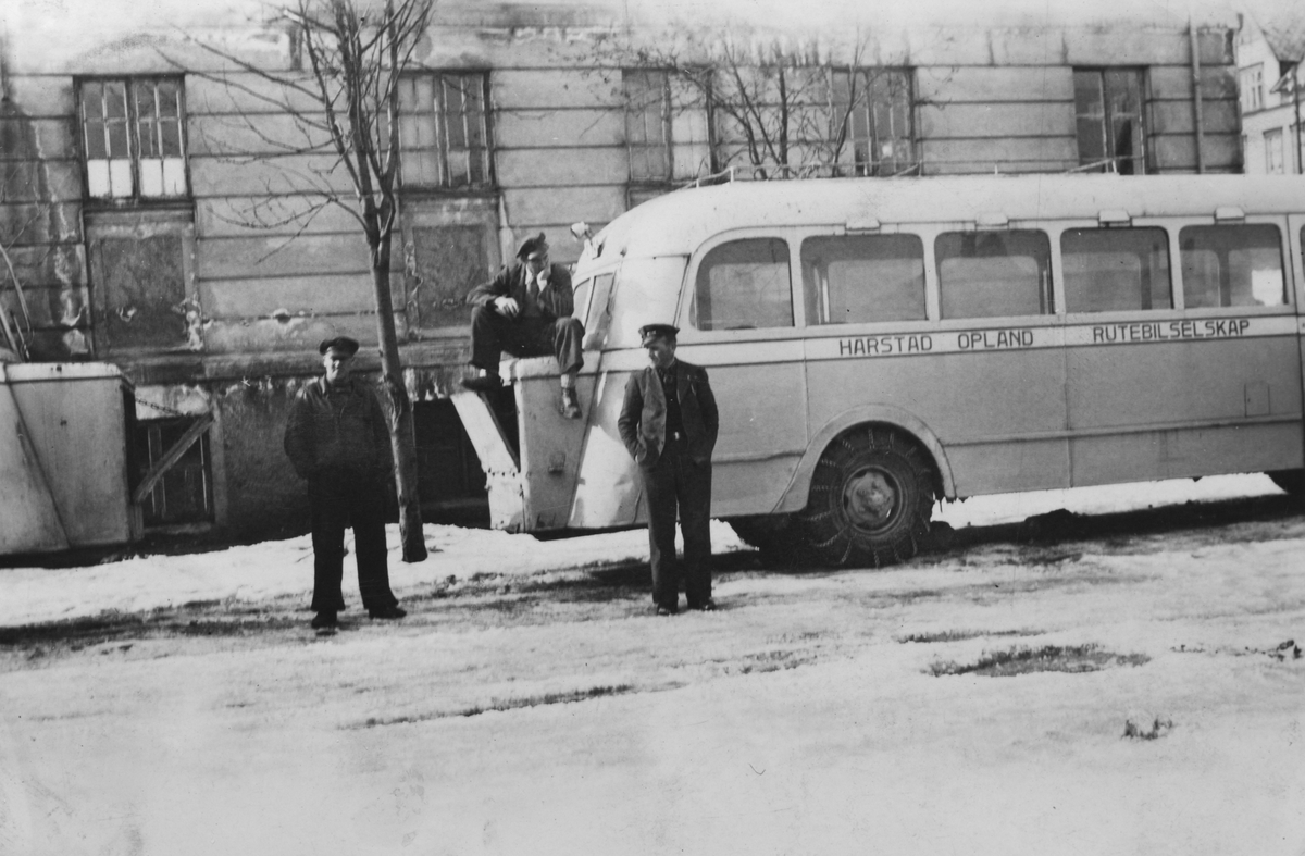 Tre menn ved en HORB-buss, parkert foran en stor murbygning. Det er vinter og bussen har kjettinger på.