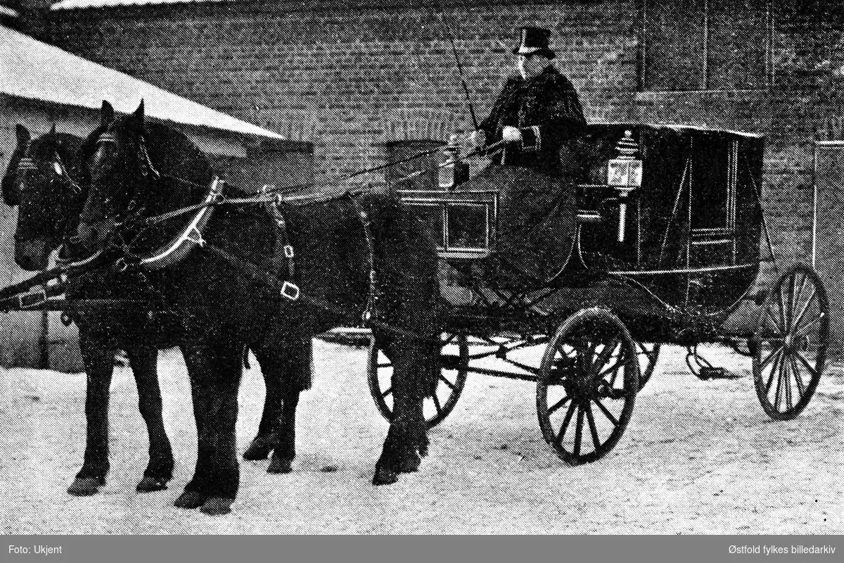 Stasvog med to hester og kusk.
Karl Edvardsens vognmannsforretning i Sarpsborg, ca. 1907-10.
Karl Edvardsen begynte som vognmann i Jernbanegta i 1896. Siden 1898 drev han med handel med meieriprodukter, særlig melk og ost, i tillegg til vedforretning.