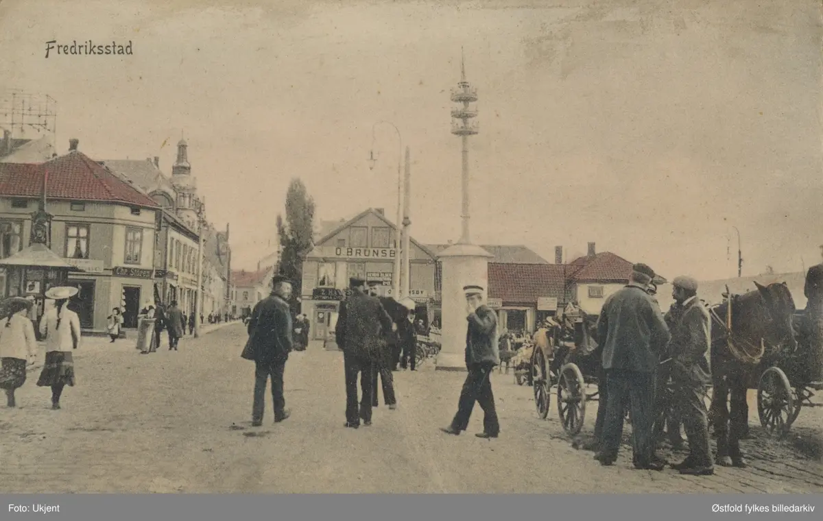Torvet. på Vestsiden i Fredrikstad. Postkort datert  1906, poststempel 1913. Postkort - lystrykk som er kolorert.
Forretningene: O. Brunsby, Chr. Støren i Storgata.