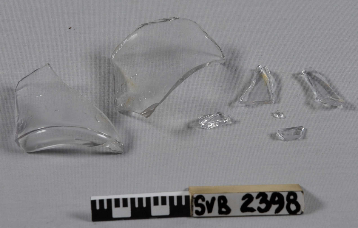 Fragmenter av glassflaske i klart glass. 2 store fragmenter og en pose med 5 mindre fragmenter. Uthevet skrift nederst på flasken.