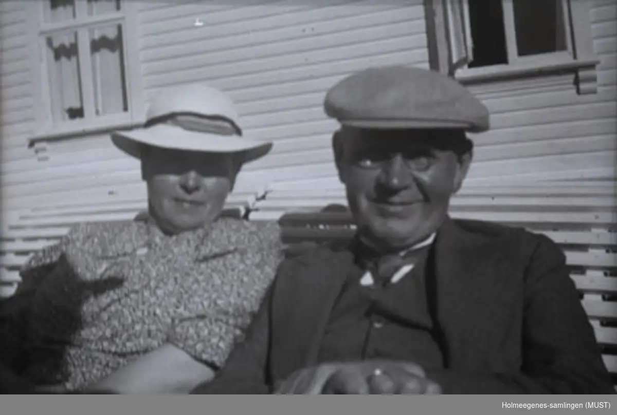 En kvinne og en mann sitter på en hagebenk foran et bolighus. Begge har hatt på. Se også ST.K.HE 2007-011-0108