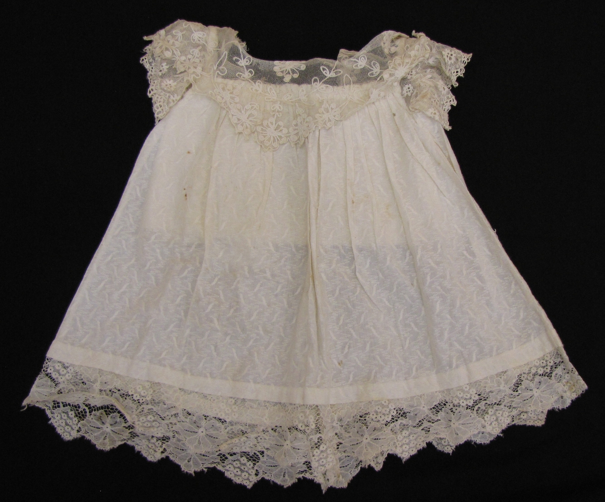 Barnklänning i vitt mönstervävt bomullstyg.

Spetsok, spetsärmar och spets nedtill på
 kjolen.