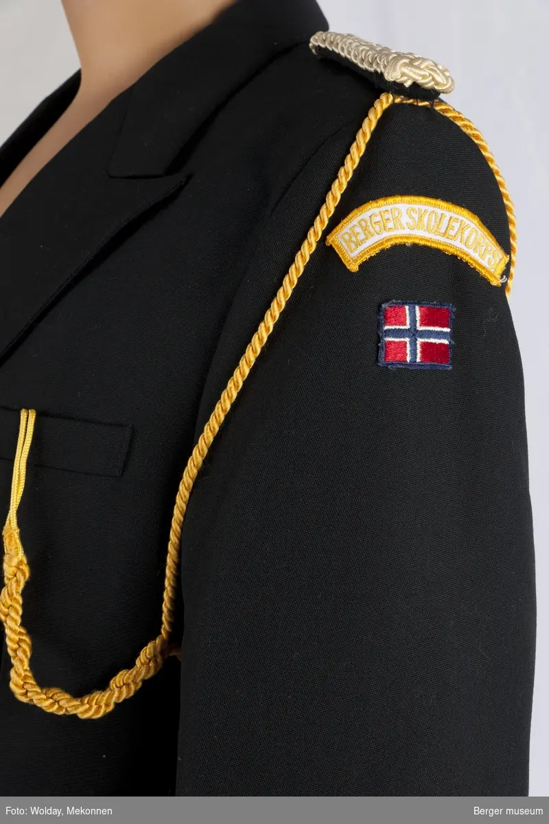 Dobbeltkneppet korpsuniformsjakke i svartt stoff med silkefor i svart. Det er pynteelementer i gull som tvunnet snor, metallknapper og epåletter i flettet snor. Venstre arm har påsydd norsk flagg.