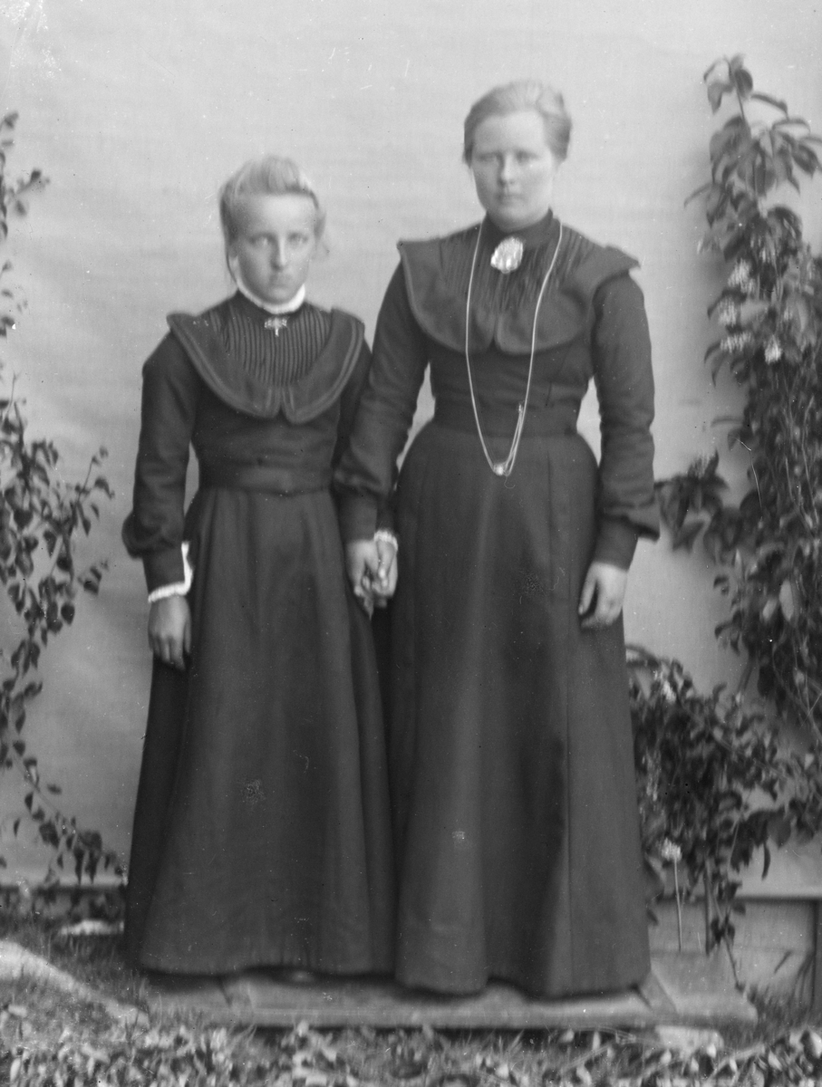 Voksen kvinne og ung jente kledd i mørke kjoler. Holder hverandre i hånda og fotografert i helfigur, med bjørkekvister foran hvitt lerret på husvegg 