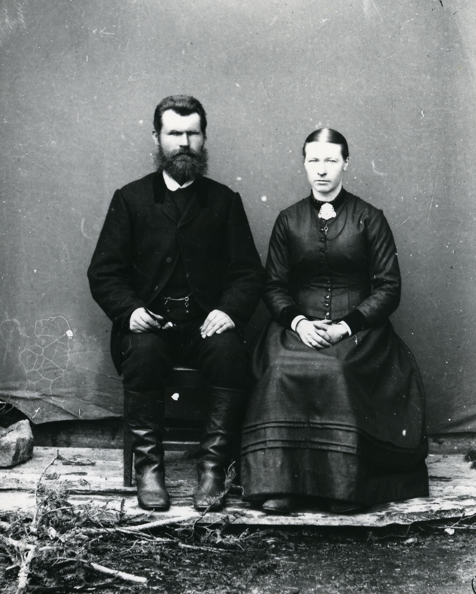 Mann i dress og skinnstøvler, og kvinne kledd i mørk kjole, sittende foran lerret