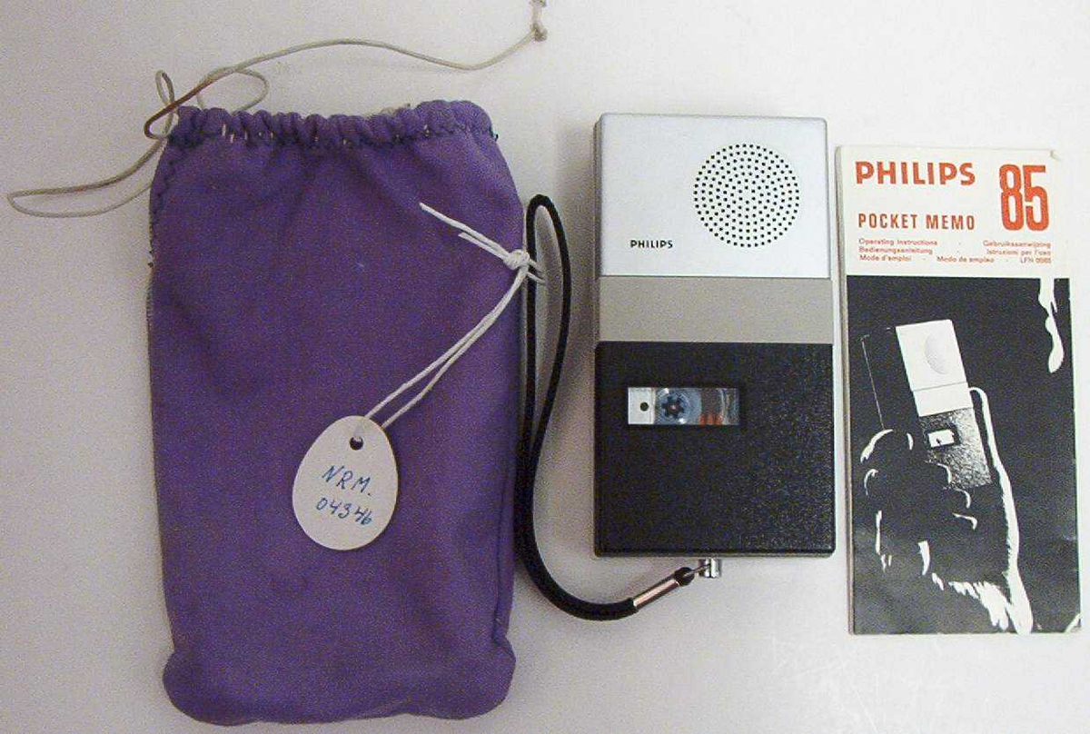 Sort, hvit og grå lommediktafon med kassett. Bruksanvisning følger med. Oppbevart i en hjemmesydd, lilla tøypose med snøring.