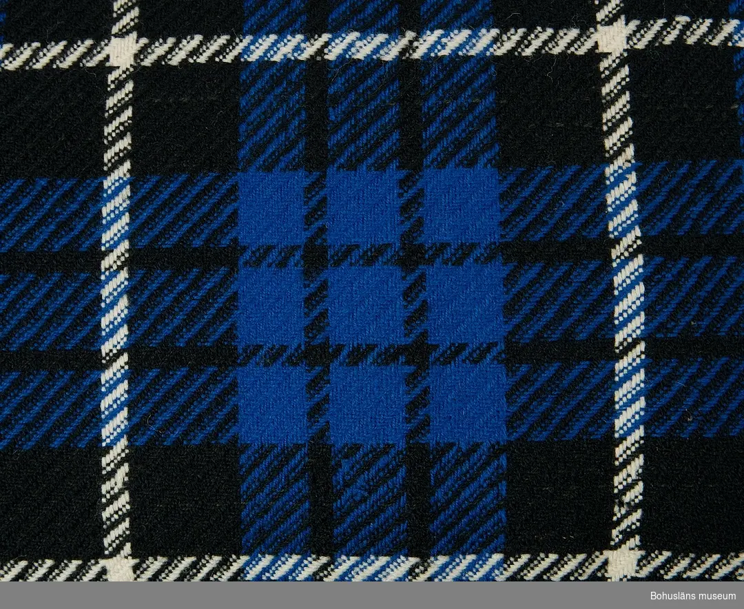 Närmast kvadratisk rutvävd ylleschal i kypert i svart, blått och vitt. Fransar runtom av varp och inslag.  Sannolikt hemvävd.