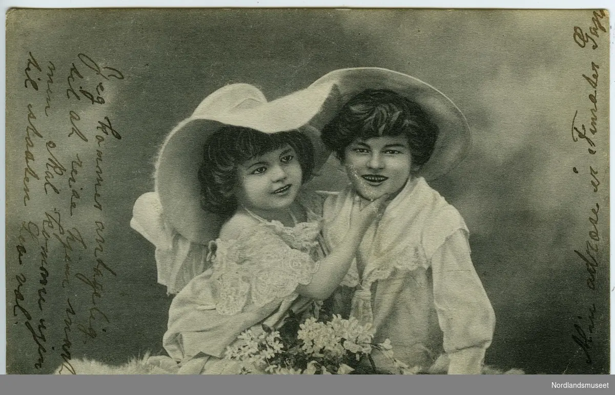 Postkort med fotografi av to småbarn. Begge har store, hvite hatter, fine klær og sitter sammen på en skinnfell el.l. Foran dem er det en blomsterbukett med hvite blomster. 

Bakside: Postfrimerke, grønt, posthorn, 5 øre. Poststemplet i Harstad 05.07.1906.